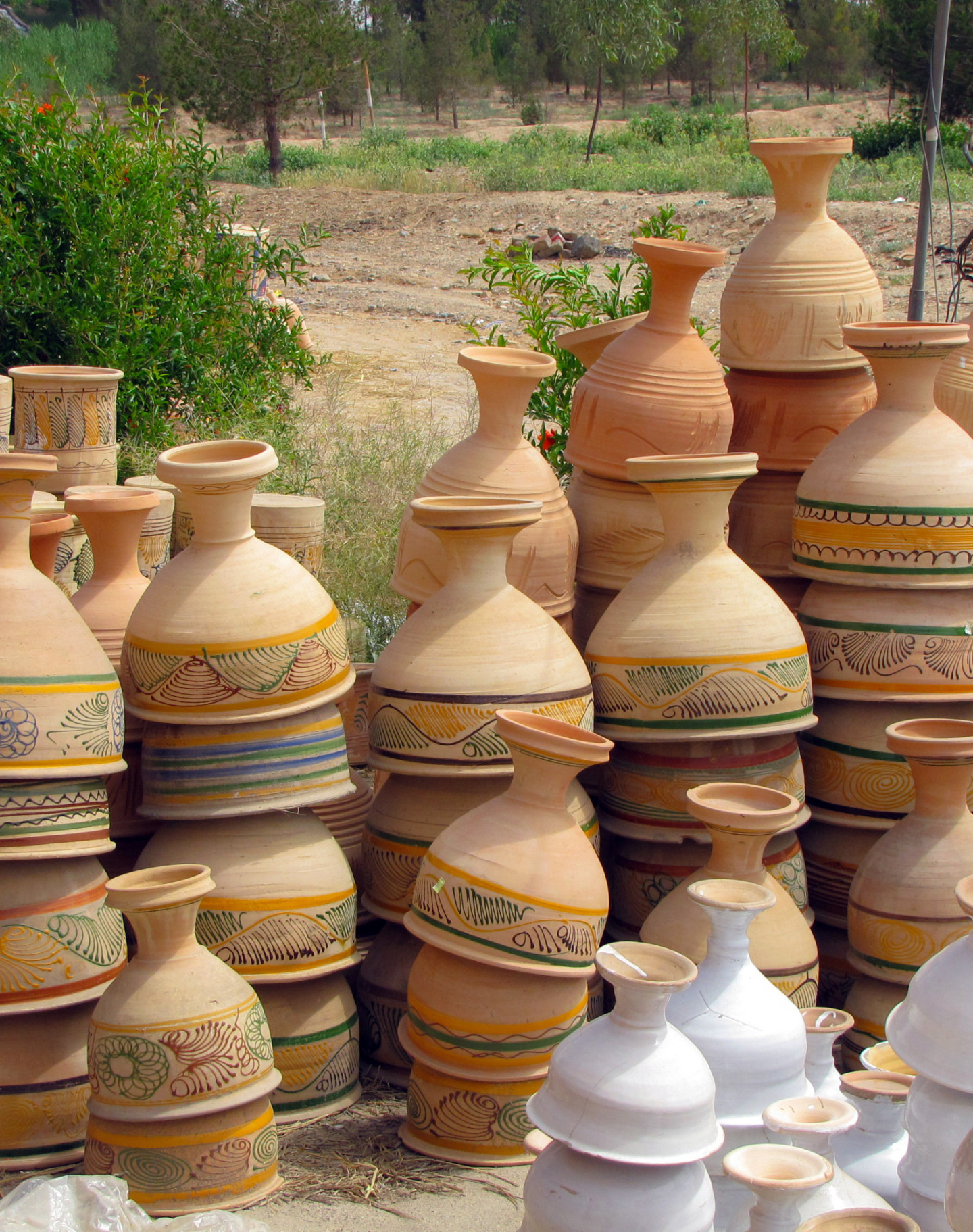 Stacked pottery, some painted white, other pieces decorated in patterns. Behind a dirt path a cluster of trees
