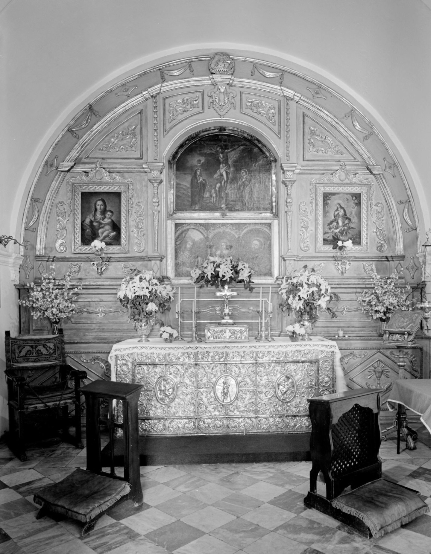 A black and white photo of an altar in an arced alcove with ornate carvings and paintings of the Christ in life, the Virgin Mary and the Cruxifiction.